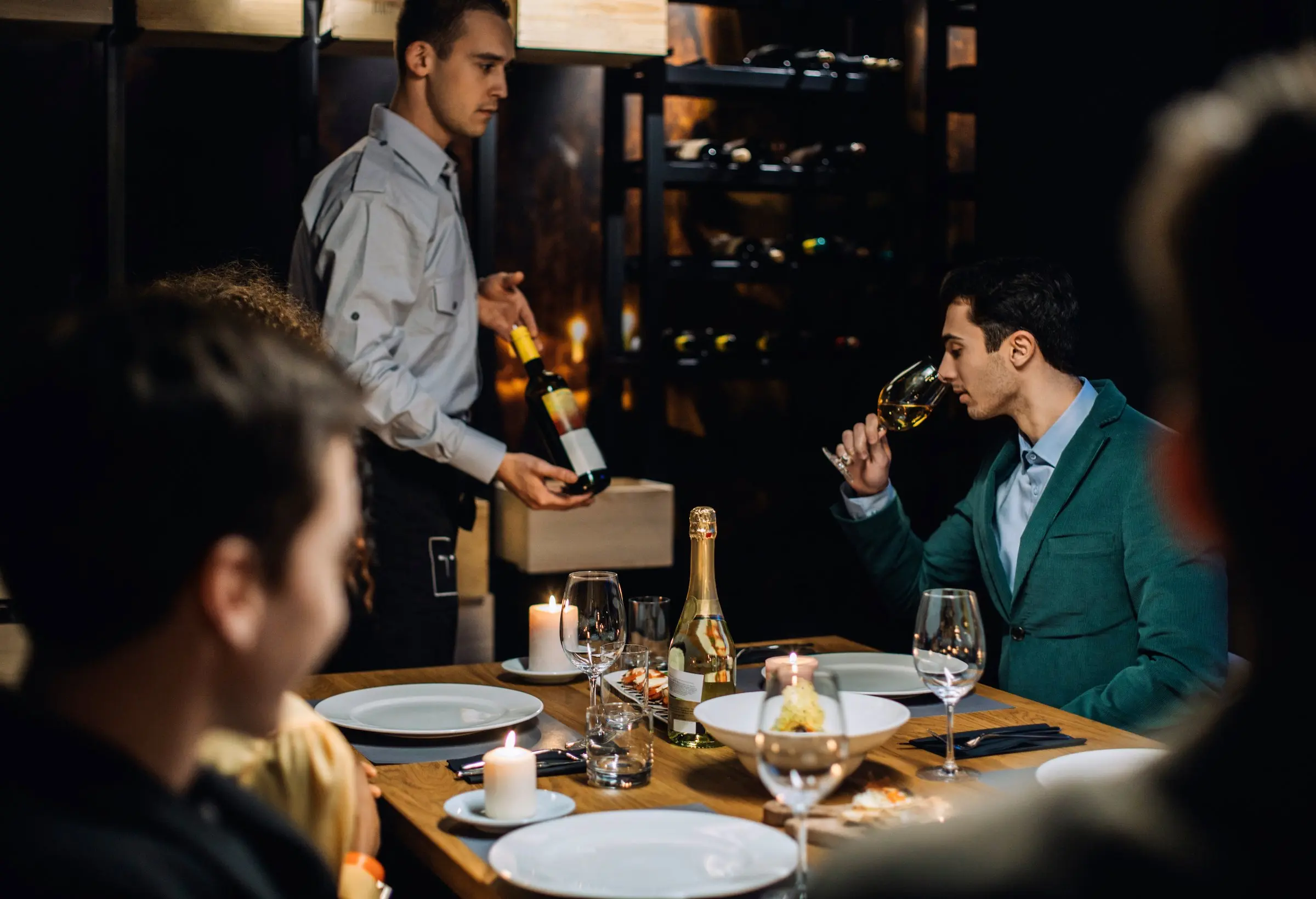 A man smelling a glass of wine at a table while other watch at Black Desert